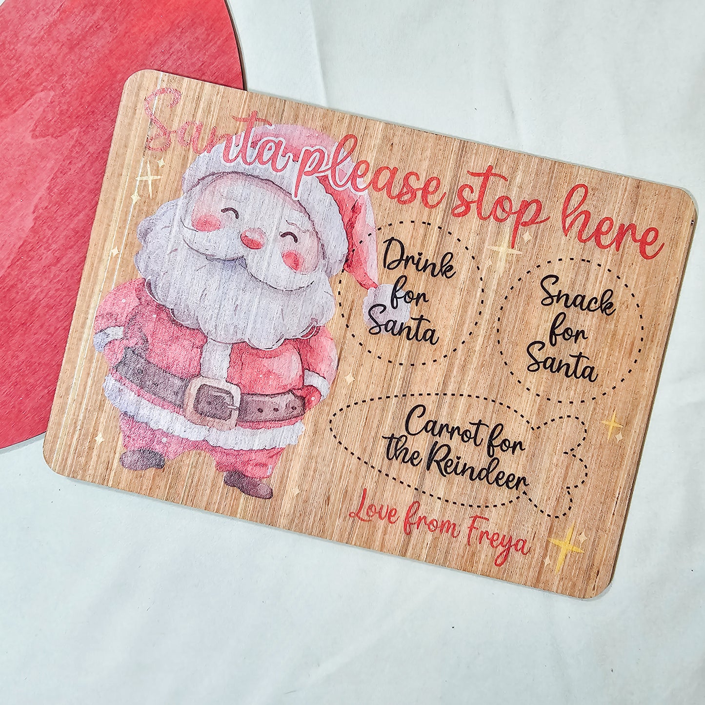 A personalised wooden Santa treat board with engraved spaces for a drink, snack, and reindeer carrot. Available in light or dark wood, with a festive Santa design and personalised name(s) at the bottom.