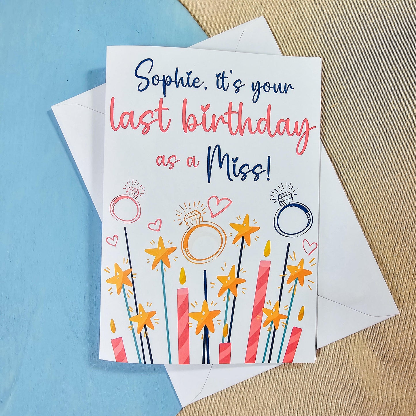A white card and envelope with 'it's your last birthday as a miss!' printed on, with rings and candles and hearts