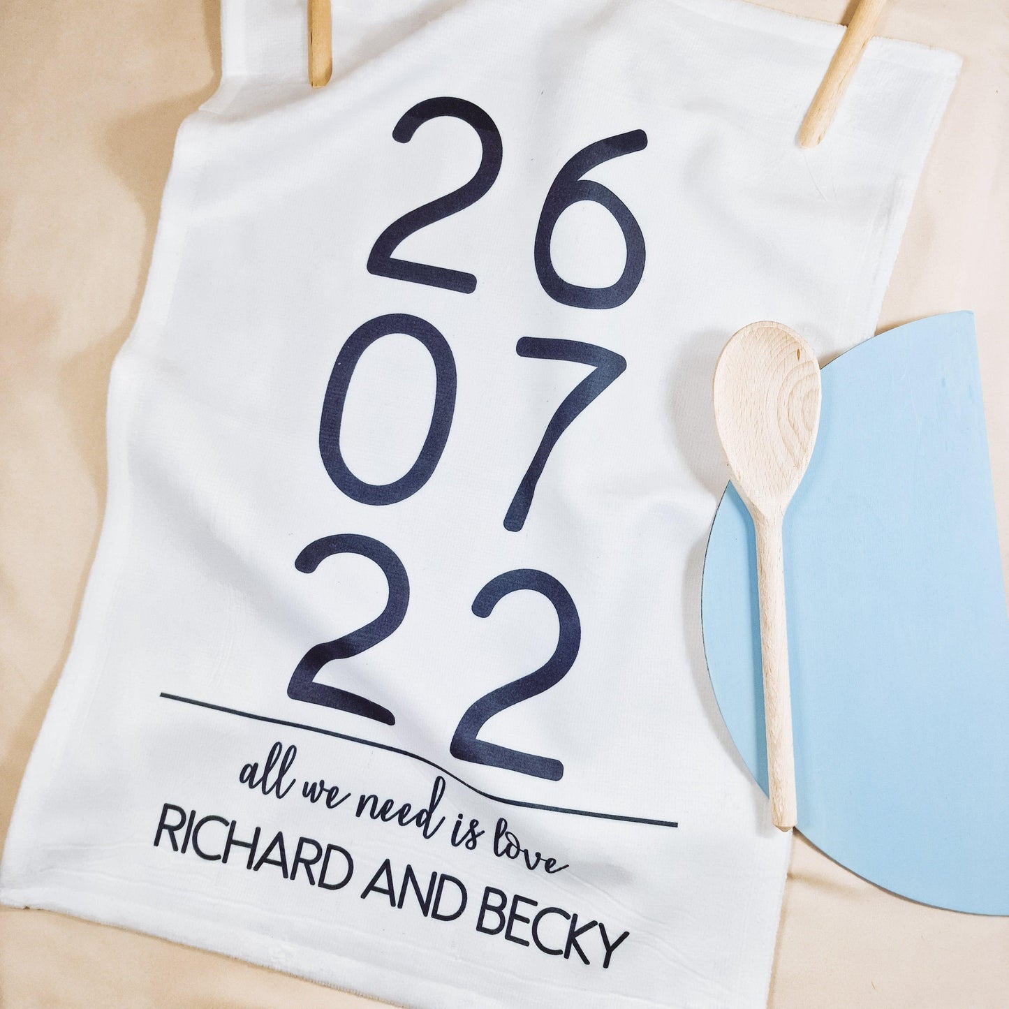 A white tea towel with the phrase "All You Need Is Love," the couple's wedding date, and their names below. The front is microfibre, and the back is 100% cotton, measuring 40x60cm. Perfect for weddings or anniversaries.