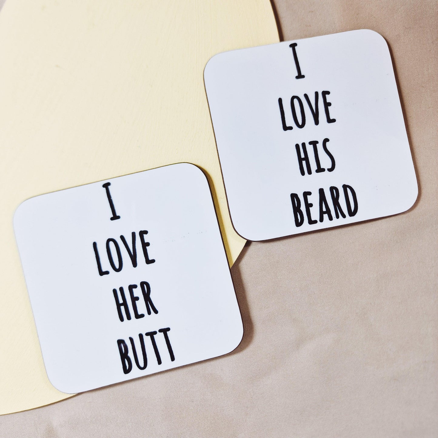 A pair of square coasters with curved edges, one saying "I Love His Beard" and the other "I Love Her Butt". Made from hardboard with a glossy, wipe-clean surface, each coaster measures approximately 9cm. Ideal for couple gifts or humorous home decor.