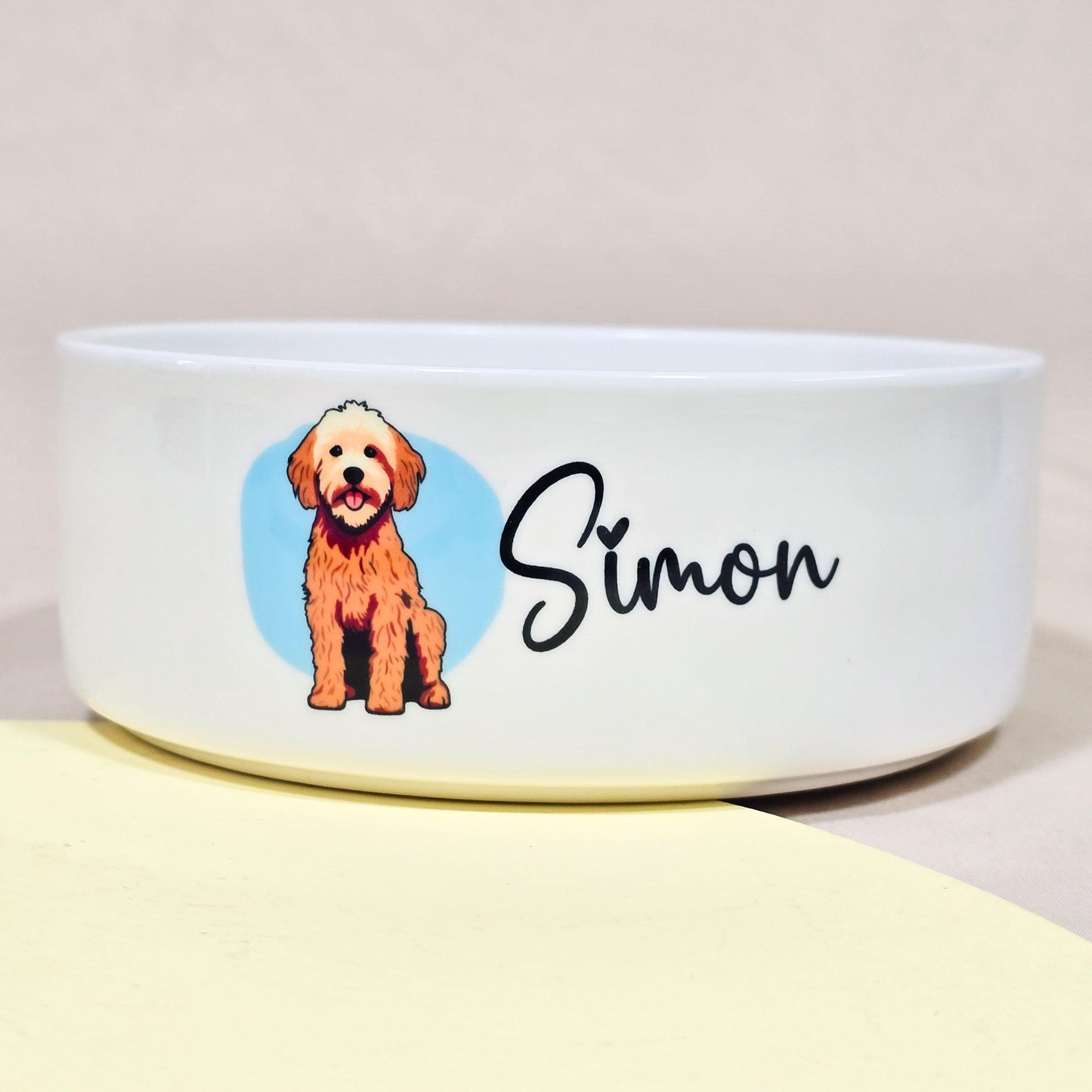 A personalised ceramic dog bowl featuring a Cockerpoo cartoon on a blue background, with the dog's name printed on it. The bowl is designed for pets and is suitable for food or water.