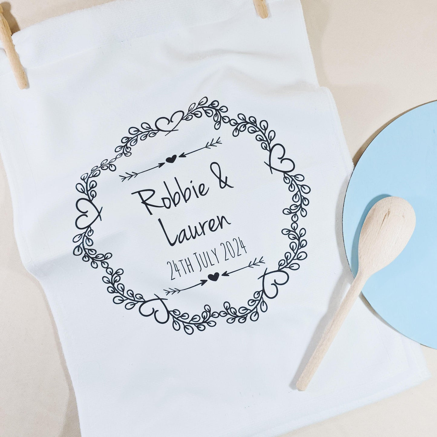A white tea towel with a circular heart and floral design, featuring the couple’s names and wedding date in the centre. Microfibre front, cotton back, measures 40x60cm, ideal for weddings or anniversaries.