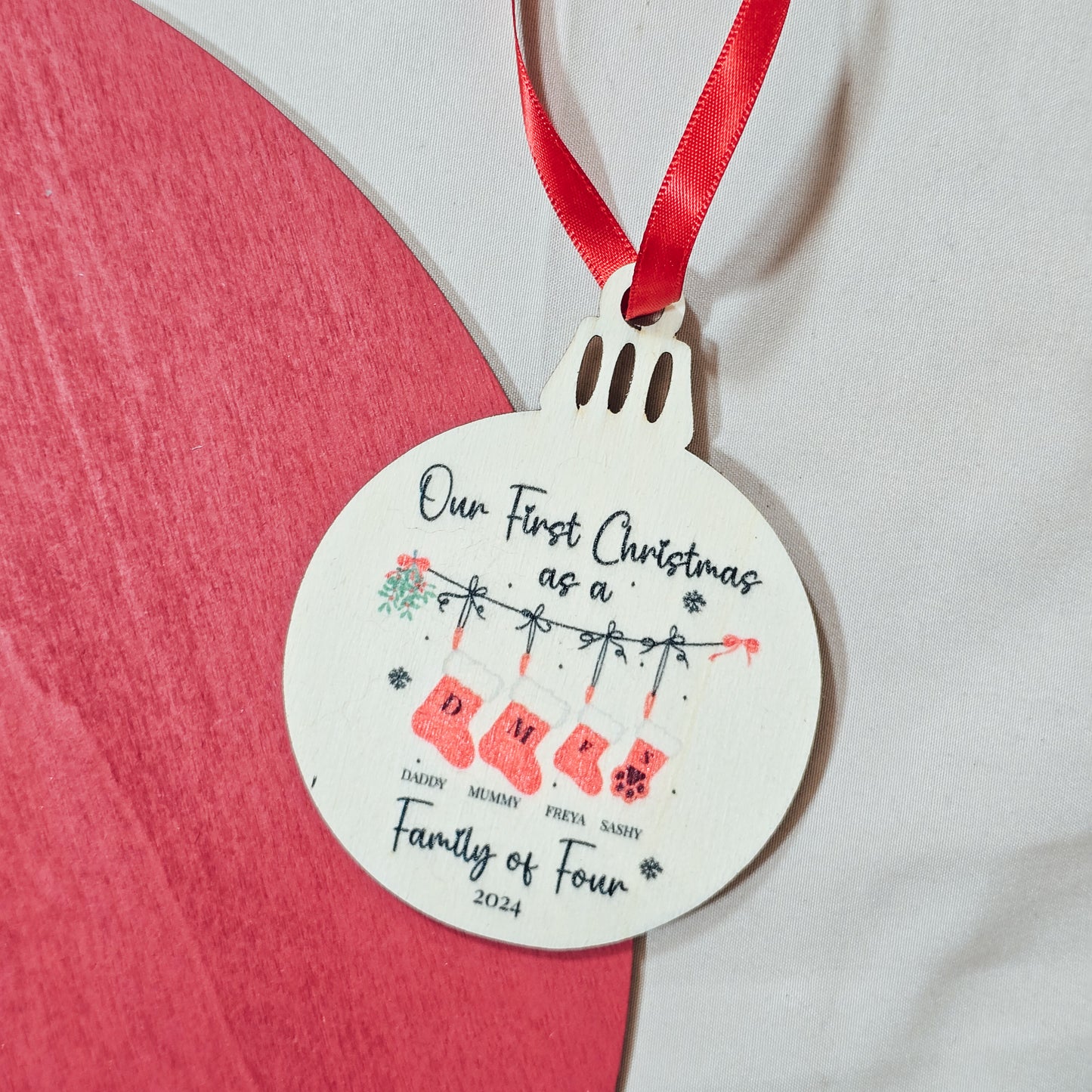 Personalised wooden bauble with "First Christmas as a Family of Four" text, featuring family members' names and stockings, including options for pets, and the year printed. Comes with ribbon for hanging.