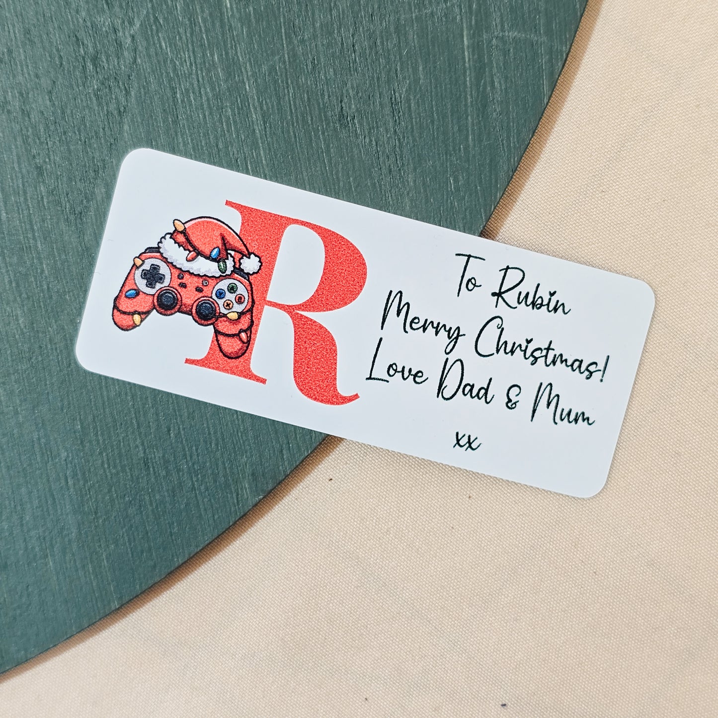 Christmas sticker featuring a gaming controller with fairy lights, a Santa hat, and a child’s initial, along with a custom festive message, in a 4cm design.