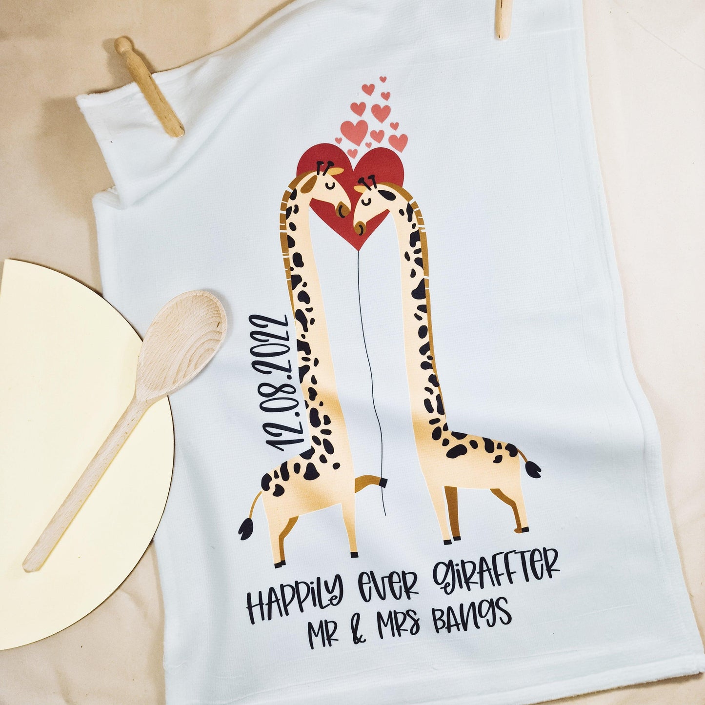A white tea towel featuring two kissing giraffes with the phrase "Happily Ever Giraffter" and personalised with the couple's names and date. The front is microfibre, and the back is 100% cotton, measuring 40x60cm. Ideal for weddings or anniversaries.