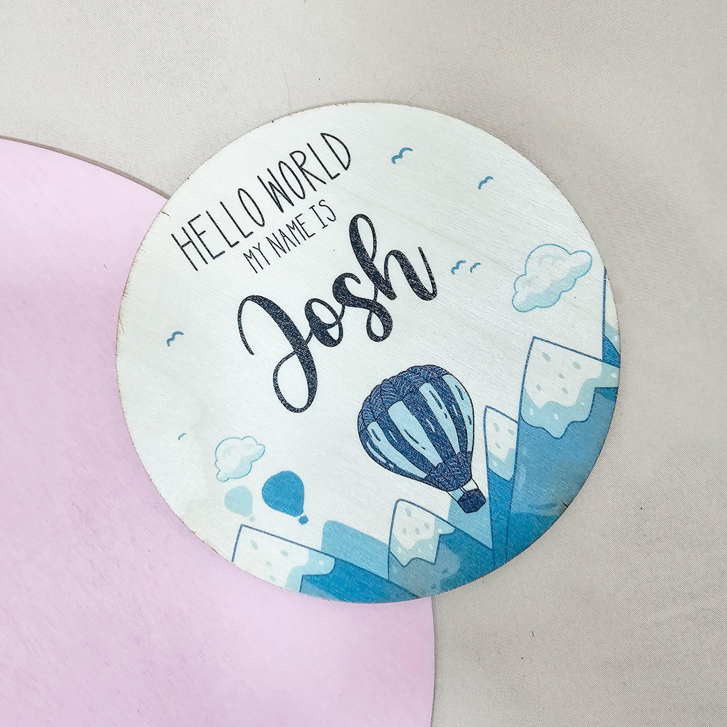 a circular wooden sign, with hot air balloons and mountains printed on and the text 'hello world, my name is'