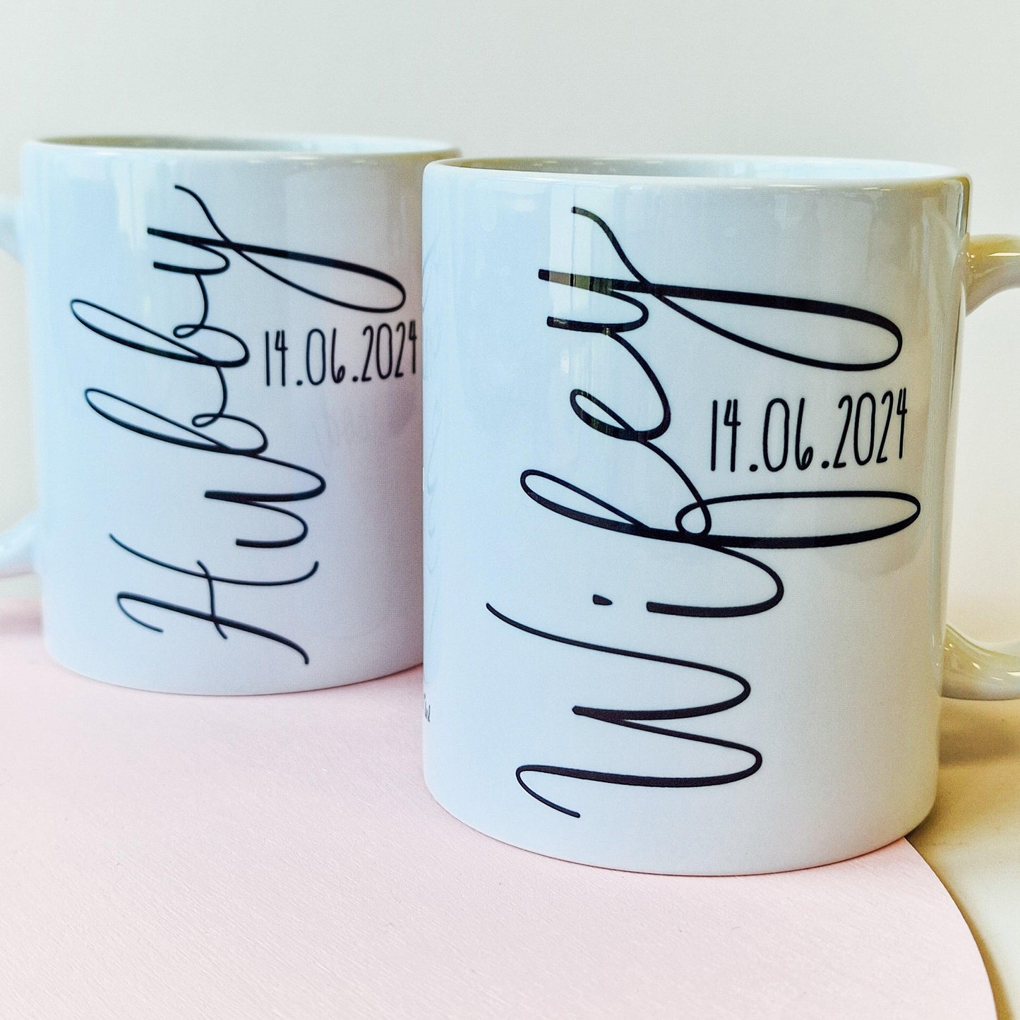 Pair of personalised mugs, one saying "Wifey" with a pink heart and wedding date, and the other saying "Hubby" with a blue heart and wedding date. Custom names on the back. Available in 10oz and 15oz sizes, dishwasher safe.