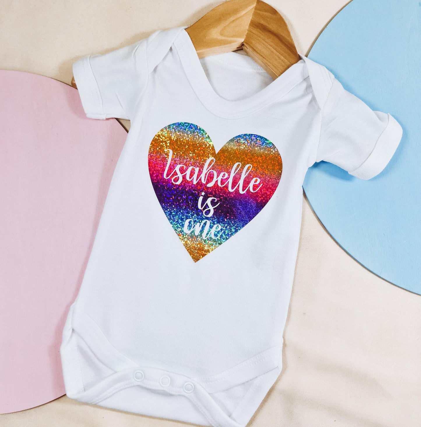 Personalised baby vest with 'Name is One' text inside a vibrant rainbow glitter heart. Perfect for celebrating a baby’s 1st birthday.