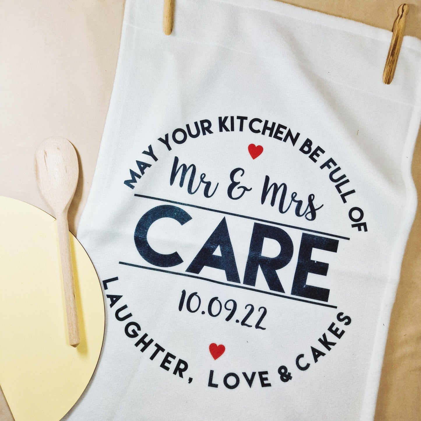 A white tea towel with the phrase "May Your Kitchen Be Full of Love, Laughter & Cakes" and the couple's name and wedding date. Microfibre front, cotton back, perfect for weddings or anniversaries, measures 40x60cm.