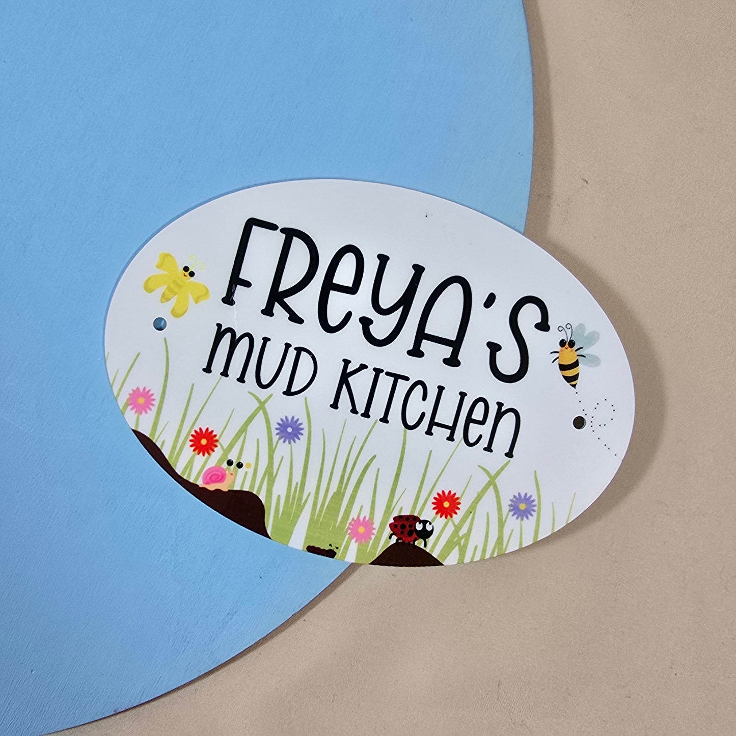 Oval-shaped sign with 'FREYA'S MUD KITCHEN' text and garden-themed illustrations on a blue and beige background.
