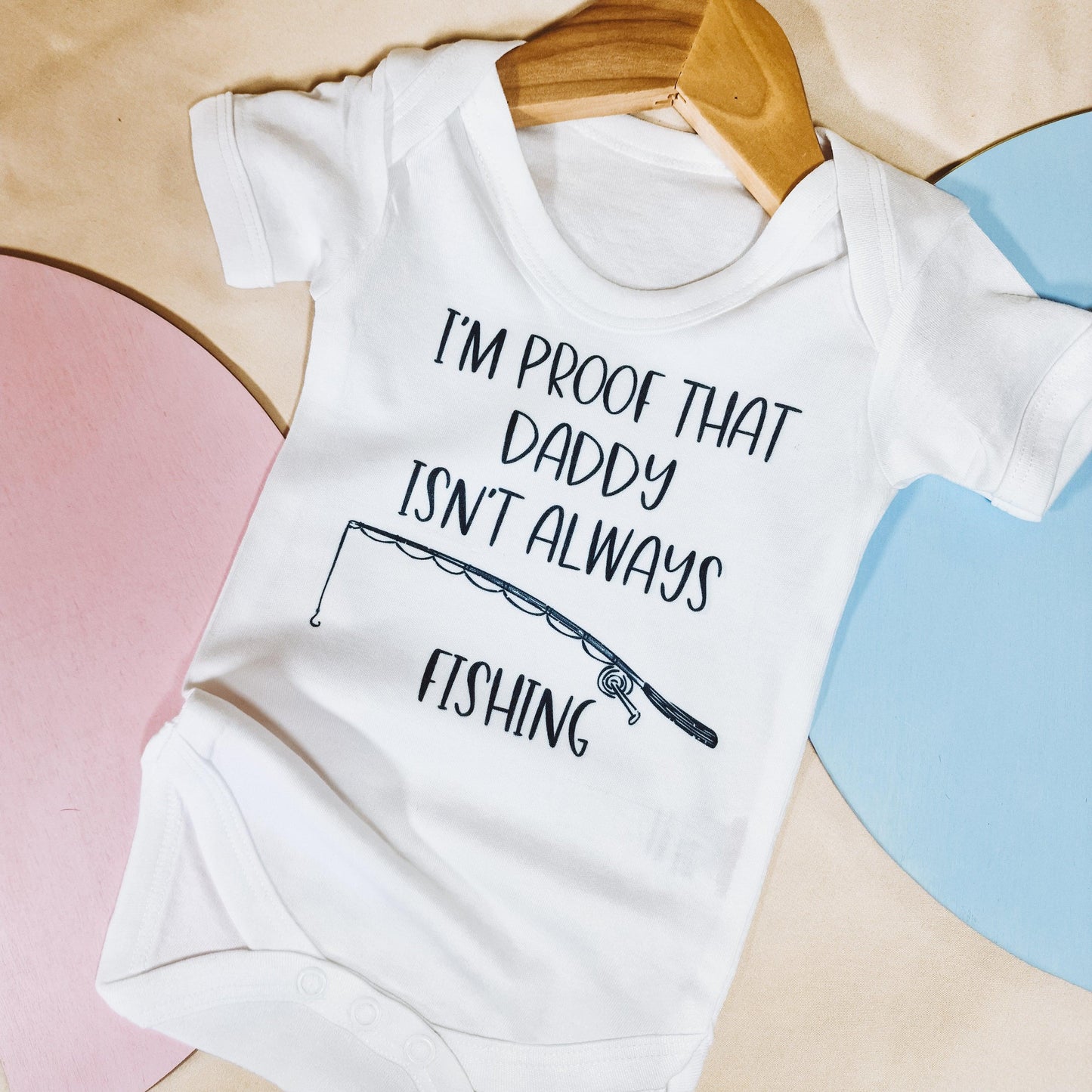 White baby vest with the text "I'm proof that my daddy isn't always fishing" and a fishing rod design, shown alongside a matching bib option. Suitable for babies from newborn to 24 months.