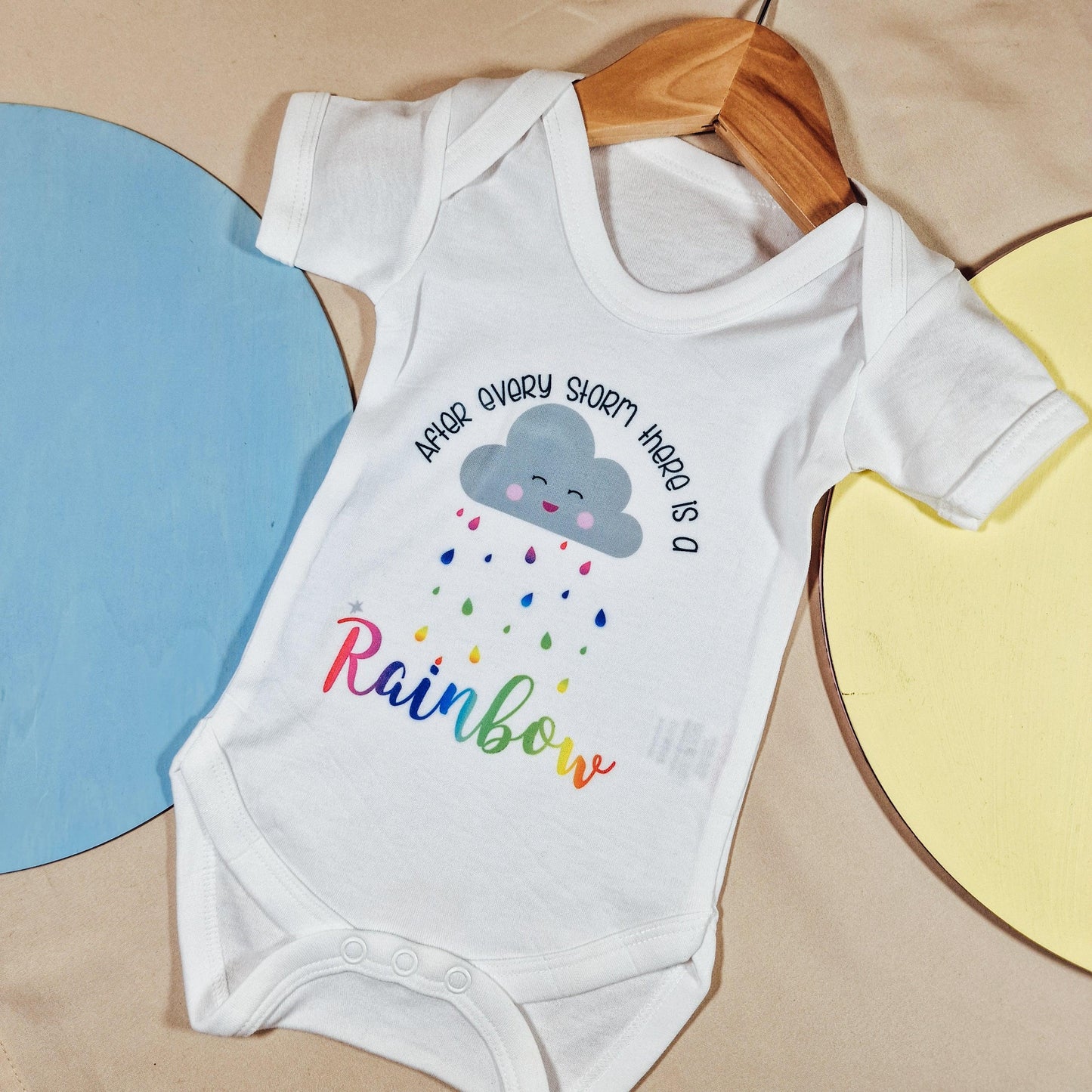 White baby vest featuring the text 'After Every Storm Is a Rainbow' in vibrant colours. Available in sizes newborn to 24 months, made from soft, machine washable fabric