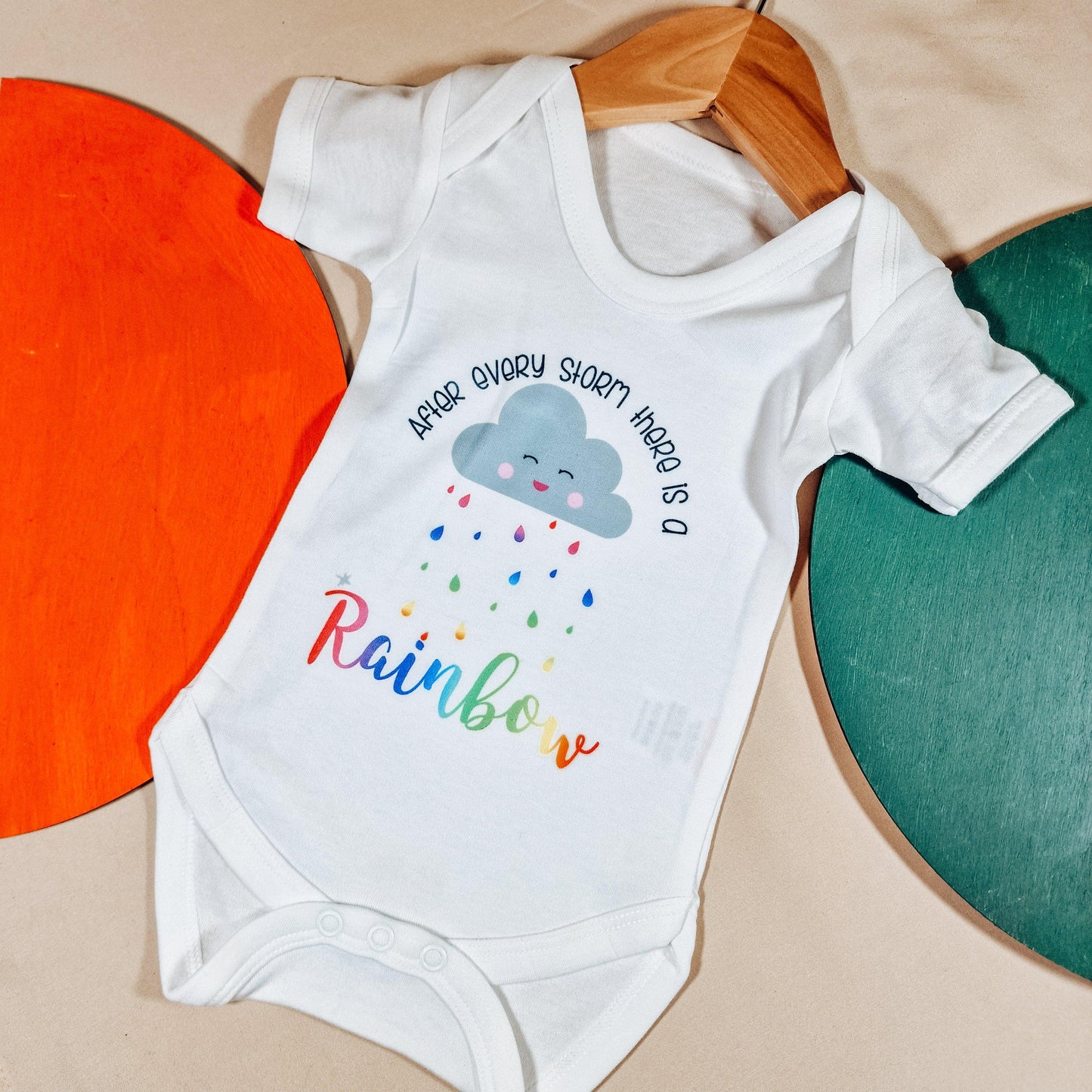 White baby vest featuring the text 'After Every Storm Is a Rainbow' in vibrant colours. Available in sizes newborn to 24 months, made from soft, machine washable fabric