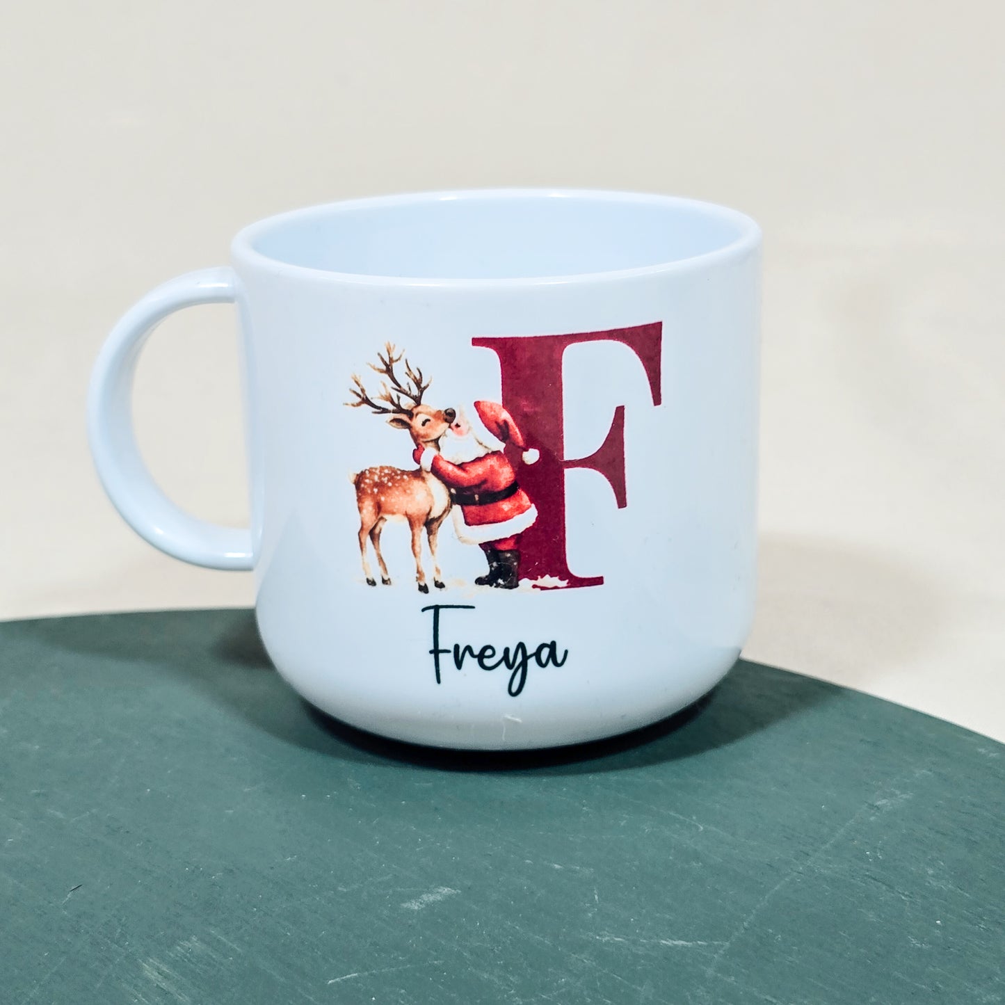 A 6oz mini children's mug featuring a festive design of Santa kissing a reindeer, personalised with a child's name and initial. The unbreakable polymer cup is perfect for little hands and makes an ideal Christmas gift, with matching adult mugs available.