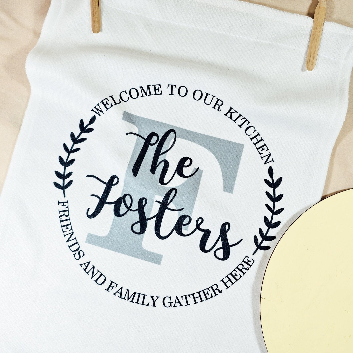 A white tea towel with a circular design that says "Welcome to Our Kitchen, Friends and Family Gather Here" and features a family name in the centre. Microfibre front, cotton back, measures 40x60cm, perfect for home decor or housewarming gifts.