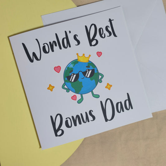 A square 15x15cm greeting card with the words "World’s Best Bonus Dad" printed on the front in bold, stylish lettering. The card is displayed alongside a white envelope on a neutral background.


