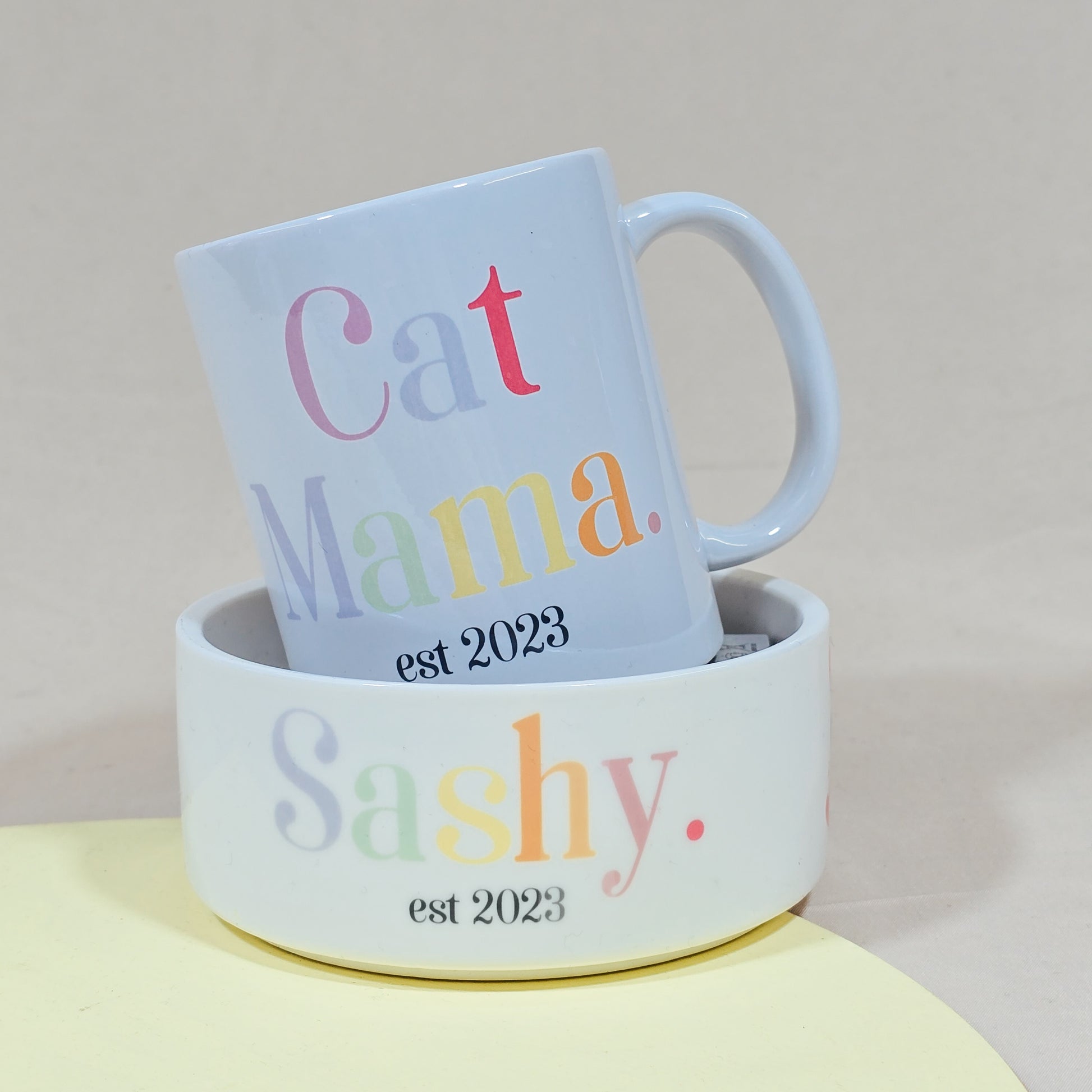 A white ceramic mug with "Cat Mama Est [Date]" in elegant script, available with a matching pet bowl in various sizes.

