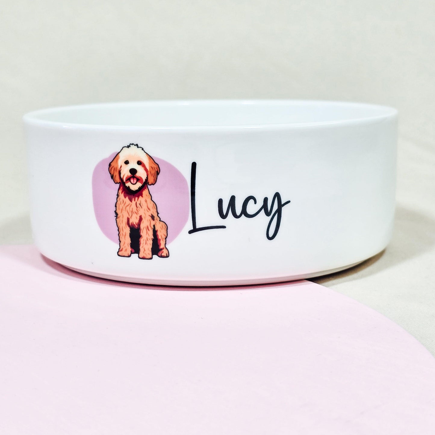 A personalised ceramic dog bowl featuring a cartoon Cockerpoo on a pink background, customisable with the dog's name. Available in small (12cm), medium (15cm), and large (18cm) sizes.