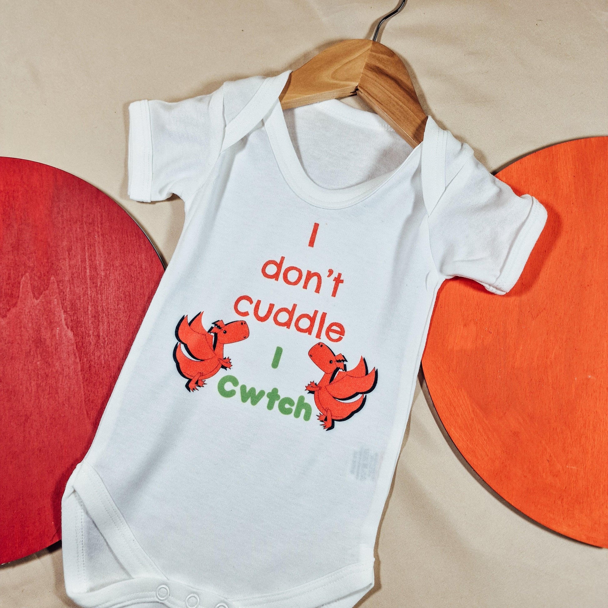 White baby vest with the text 'We Don’t Cuddle, We Cwtch' and two red Welsh dragons. Available in sizes newborn to 24 months, made from soft, machine washable fabric.