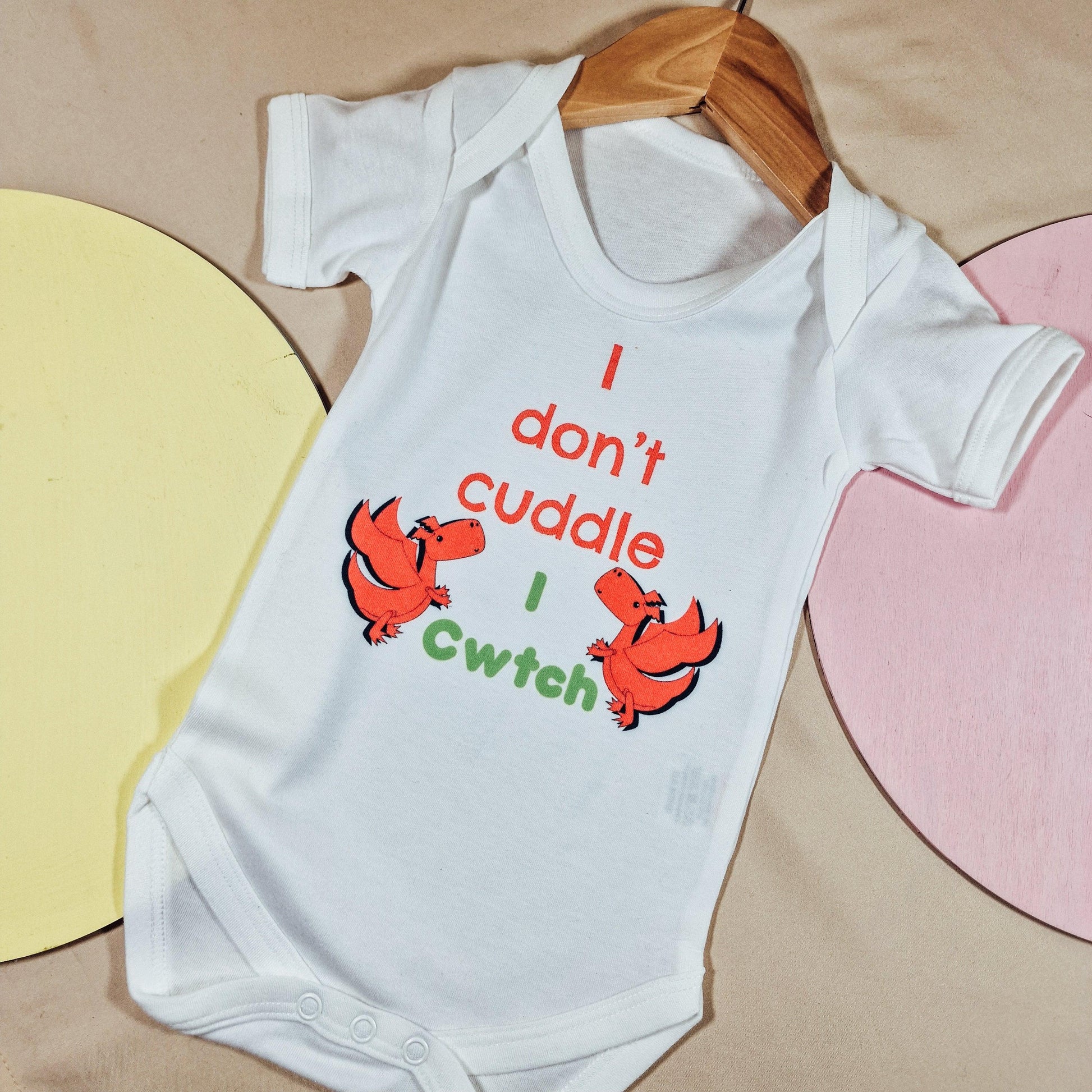 White baby vest with the text 'We Don’t Cuddle, We Cwtch' and two red Welsh dragons. Available in sizes newborn to 24 months, made from soft, machine washable fabric.