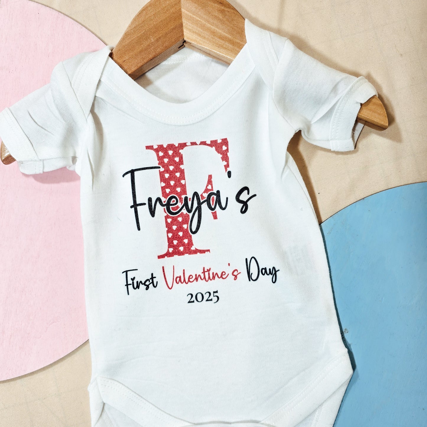 A white baby vest with a red heart-filled initial, personalised with the text "[Name’s] First Valentine’s [Year]," perfect for celebrating a baby’s first Valentine’s Day.