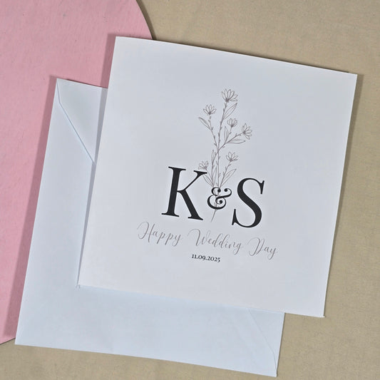 Square wedding card featuring couple’s initials, the phrase "Happy Wedding Day," wedding date, and a delicate floral design.


