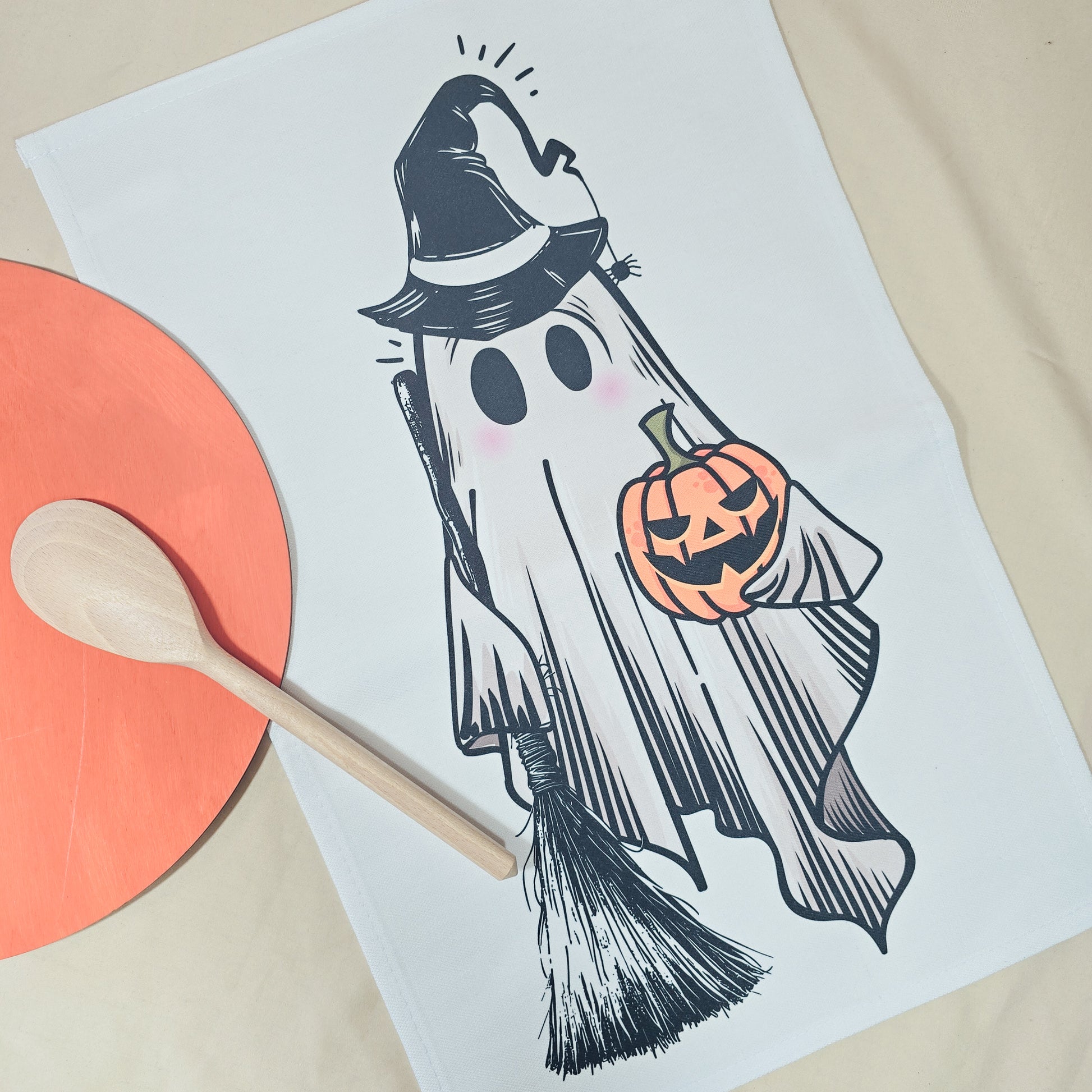 Halloween-themed ghost illustration on a tea towel with a wooden spoon and orange circular object.