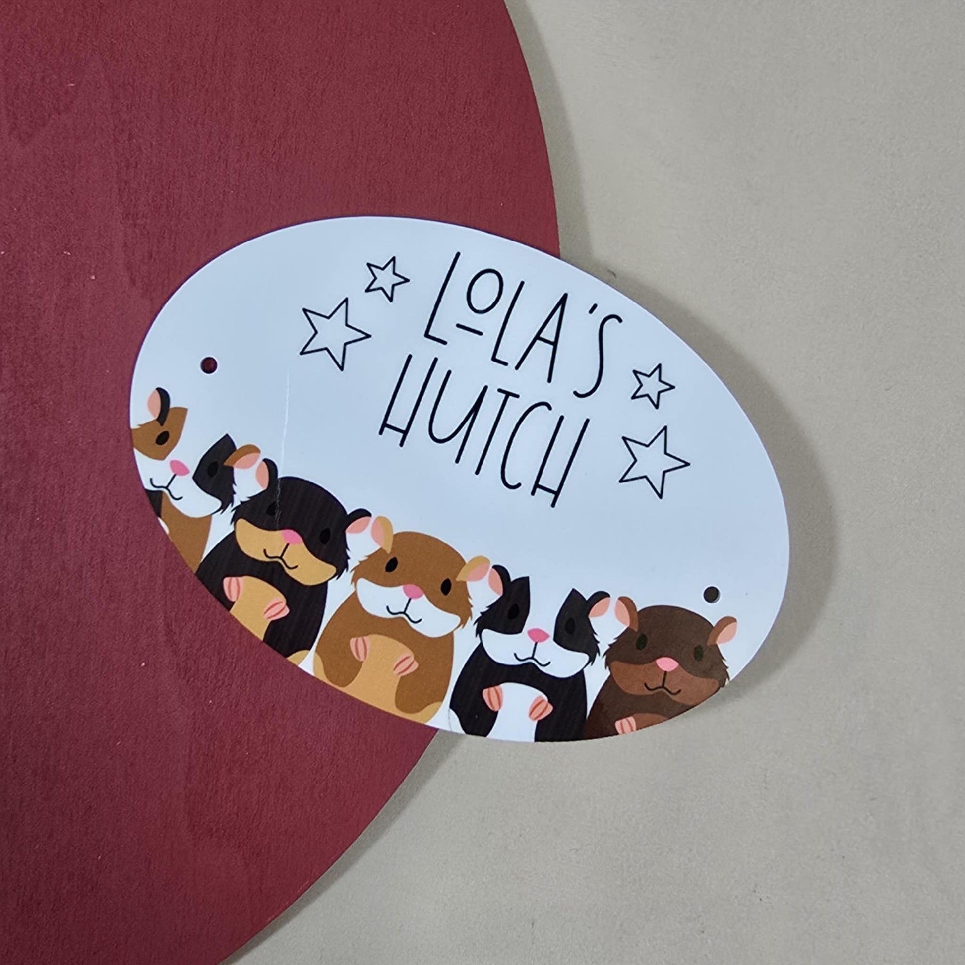 A small rectangular door plaque (approx 12.5x9cm) featuring two guinea pigs and the text “[Name]’s Hutch” with two holes for hanging. Custom text available.

