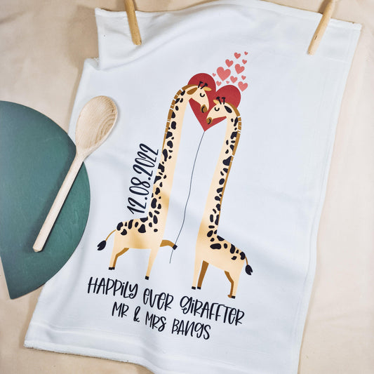 A white tea towel featuring two kissing giraffes with the phrase "Happily Ever Giraffter" and personalised with the couple's names and date. The front is microfibre, and the back is 100% cotton, measuring 40x60cm. Ideal for weddings or anniversaries.