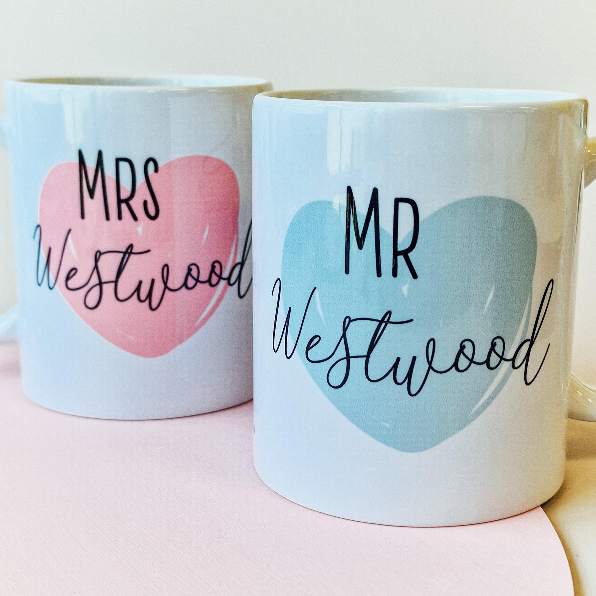 Pair of personalised mugs, one saying "Wifey" with a pink heart and wedding date, and the other saying "Hubby" with a blue heart and wedding date. Custom names on the back. Available in 10oz and 15oz sizes, dishwasher safe.