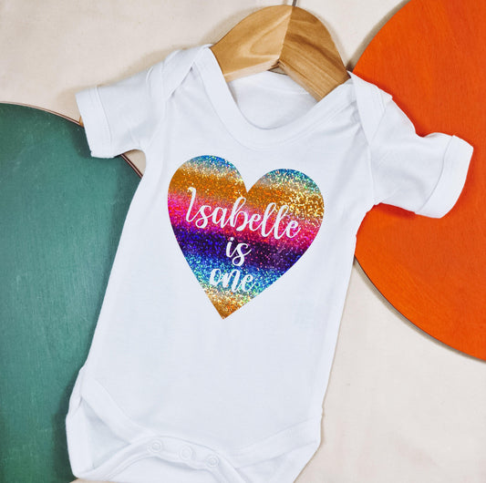 Personalised baby vest with 'Name is One' text inside a vibrant rainbow glitter heart. Perfect for celebrating a baby’s 1st birthday.