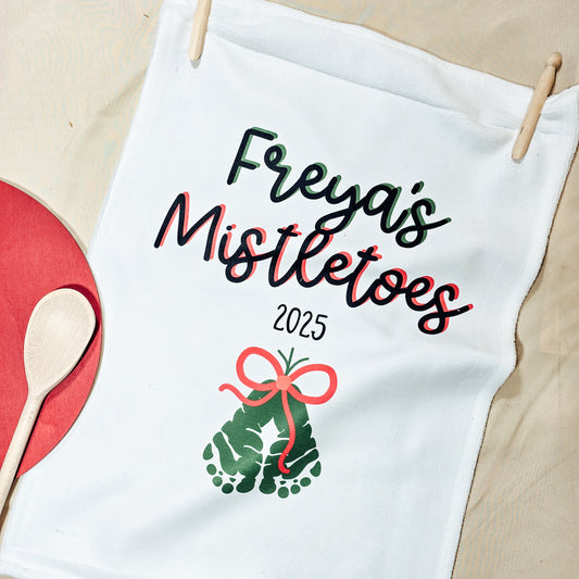 White towel with 'Freyas Mistletoes 2025' text and footprints, hanging on a wooden stick.