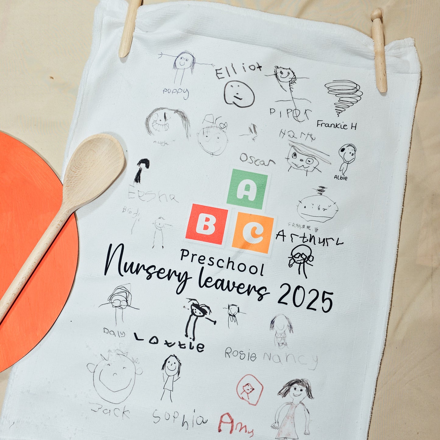 White towel with children's drawings and text, placed on a beige surface with a wooden spoon and orange circle.