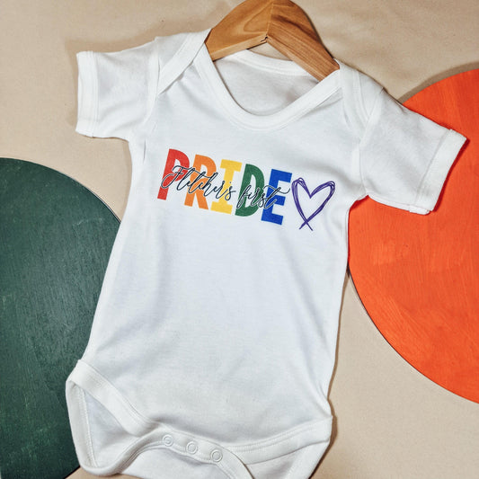 White baby vest with the personalised text '[Name]'s First Pride' and 'Pride' in rainbow colours. Available in sizes newborn to 24 months, featuring soft fabric and a machine washable design.