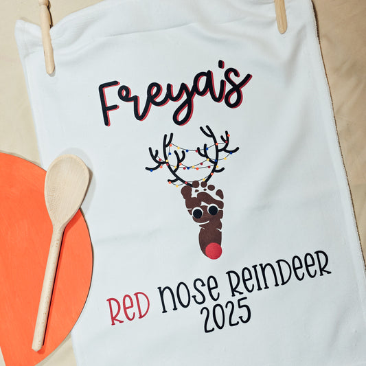 Personalised Christmas tea towel featuring a baby’s footprint turned into a red-nosed reindeer with antlers, reading “[Name]’s Red Nose Reindeer [Year]”.