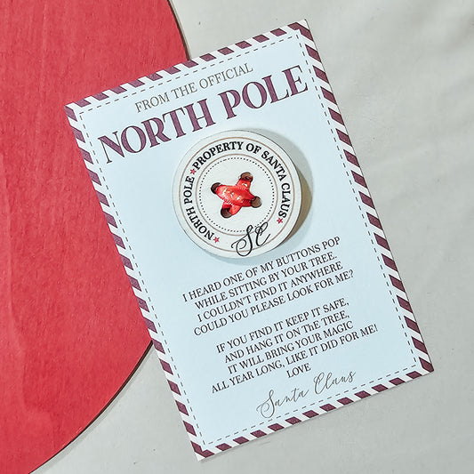 A 5cm circular button engraved with "North Pole - Property of Santa Claus," threaded with a festive ribbon that reads "Property of the North Pole." The button is accompanied by a small story card explaining how Santa lost it, adding a magical touch to Christmas morning.