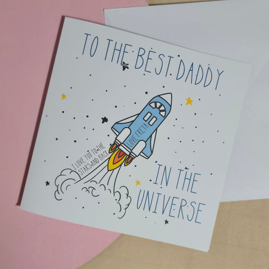 Greeting card with a rocket ship and text 'To the best daddy in the universe' on a pink background.
