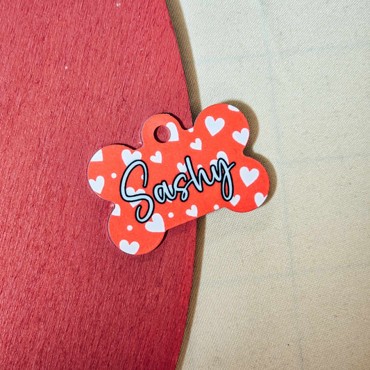 A red bone-shaped dog tag covered in love hearts, personalised with a dog’s name or initial, with owner details printed on the back, shown with a secure clip for attaching to a collar.







