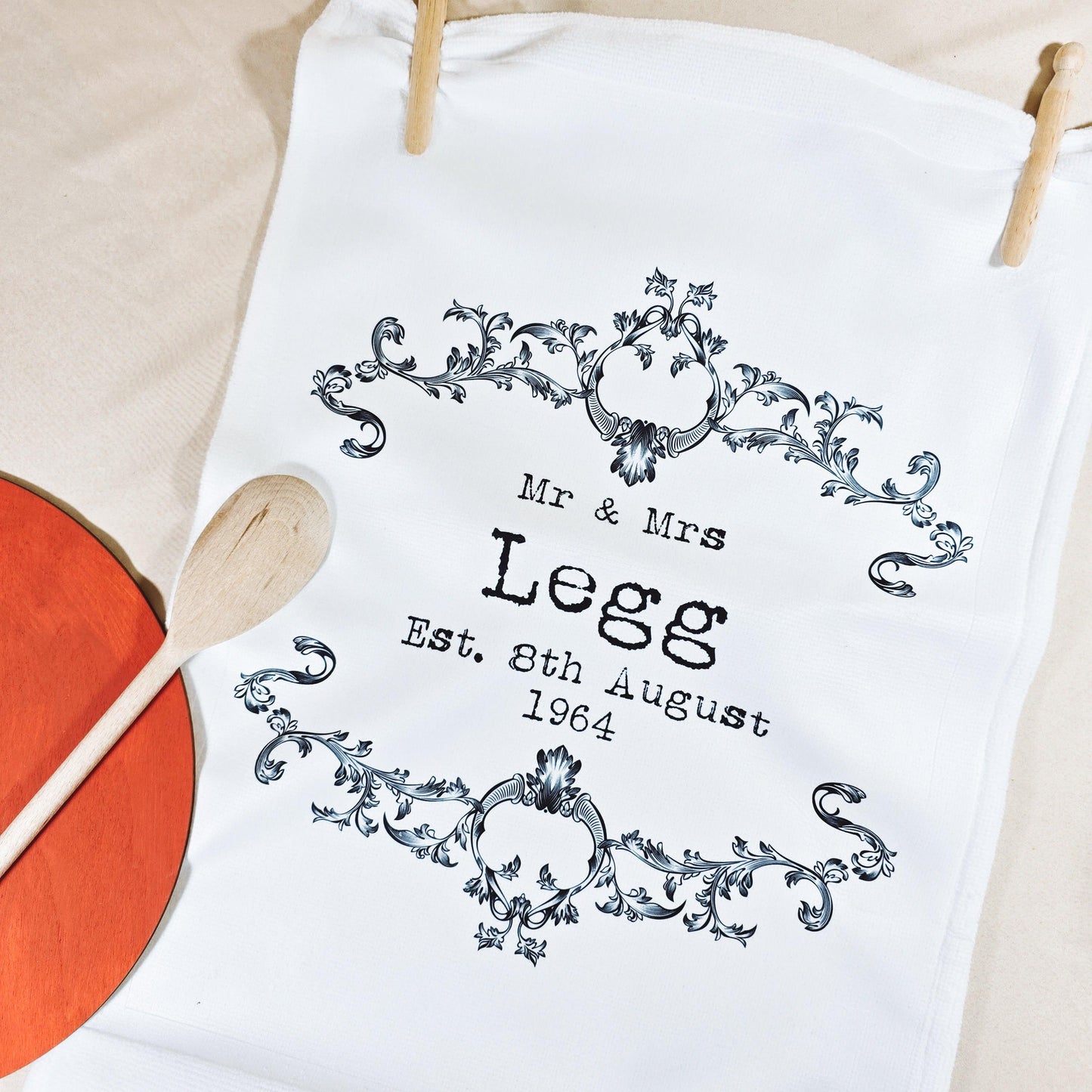 A white tea towel with a vintage-style design featuring the couple’s names and wedding date in a decorative frame. Microfibre front, cotton back, measures 40x60cm, ideal for weddings or anniversaries.