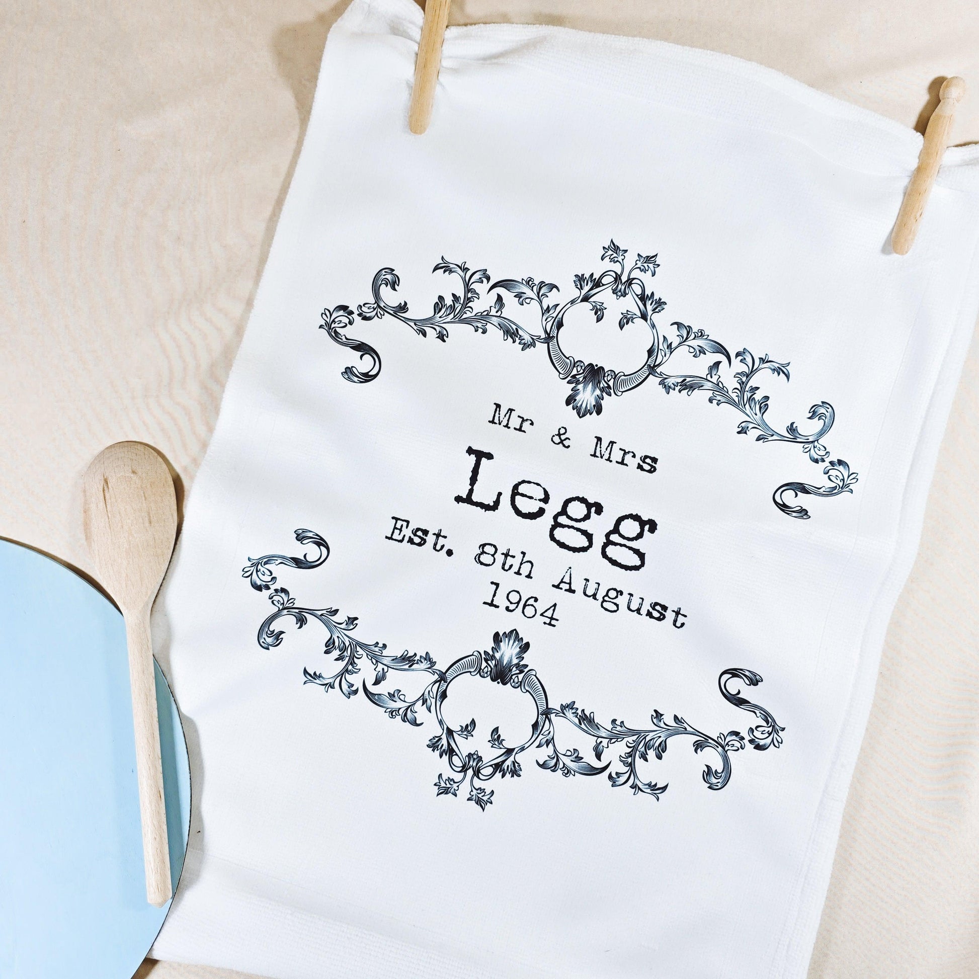 A white tea towel with a vintage-style design featuring the couple’s names and wedding date in a decorative frame. Microfibre front, cotton back, measures 40x60cm, ideal for weddings or anniversaries.