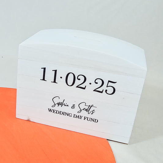 A personalised wooden moneybox, approximately 15cm wide x 11cm tall, featuring names and a wedding date with the text "Wedding Day Fund."

