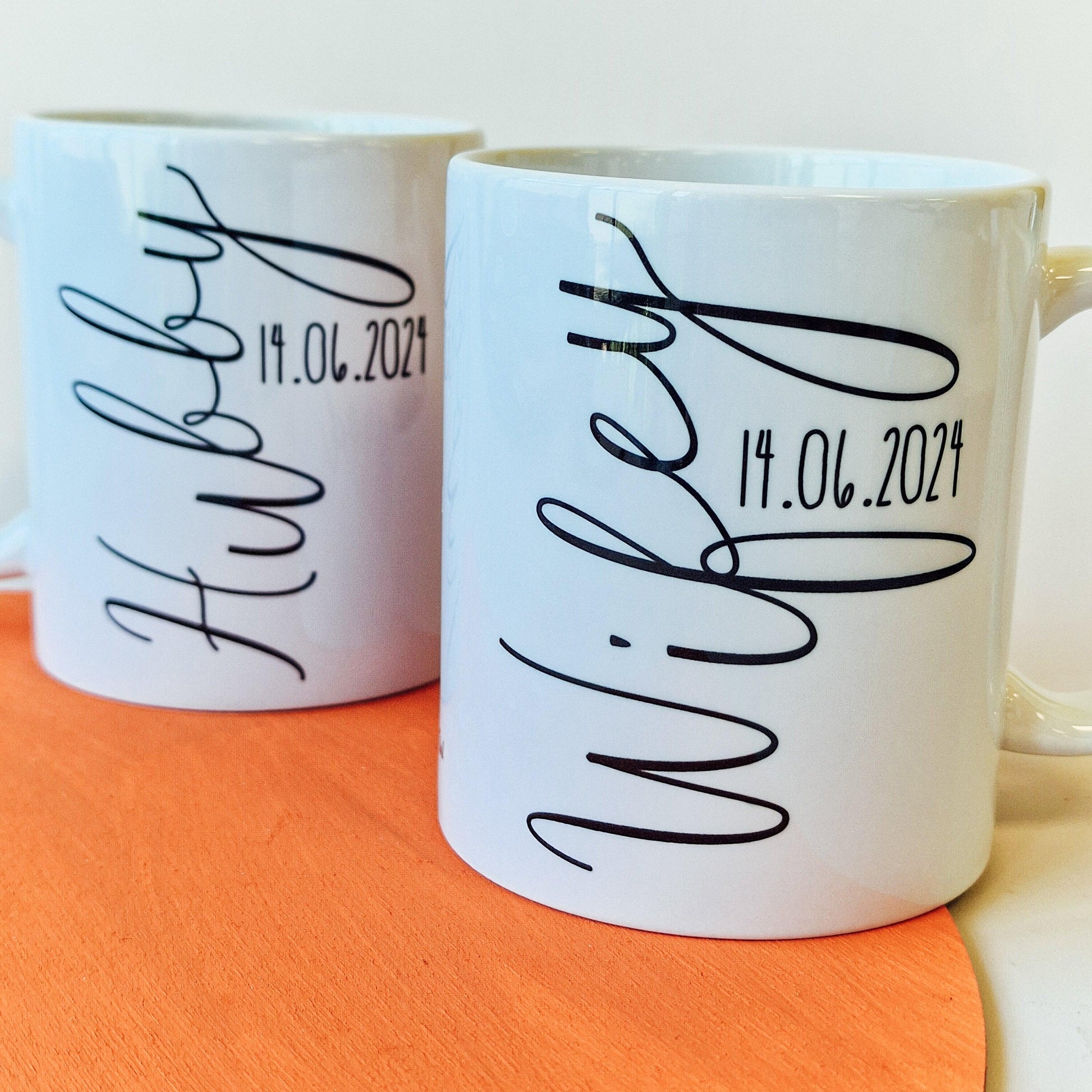 Pair of personalised mugs, one saying "Wifey" with a pink heart and wedding date, and the other saying "Hubby" with a blue heart and wedding date. Custom names on the back. Available in 10oz and 15oz sizes, dishwasher safe.